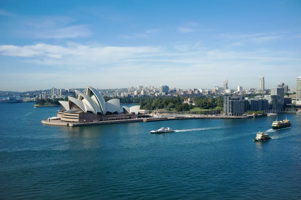 A beautiful panoramic landscape view of the Sydney Opera House and ferries on the blue waters of Sydney Harbour under a clear sky, illustrating safe Australia solo travel for women over 50.