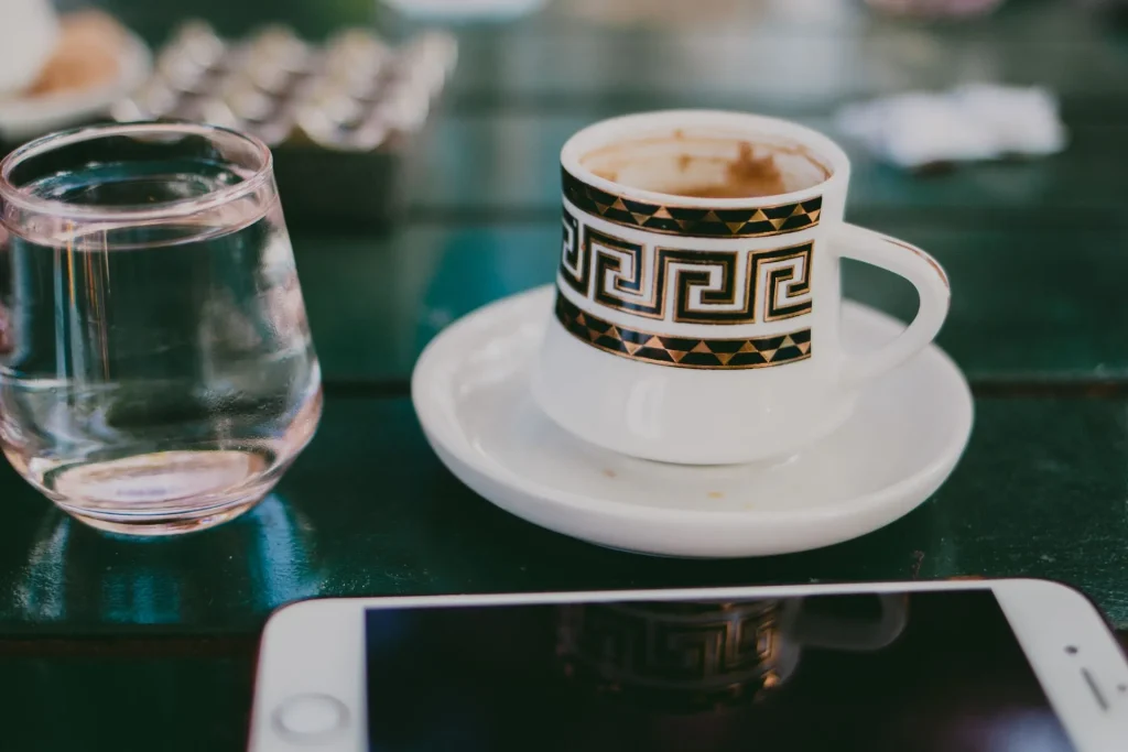 A beautiful minimalist still-life view of a traditional Greek-pattern espresso cup, a glass of water, and a smartphone resting on a café table, illustrating how easy staying connected abroad is for women over 50.