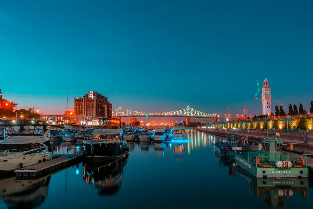 Solo travel Montreal — the Old Port at blue hour, boats moored in the marina below the illuminated Jacques Cartier Bridge and the glowing Clock Tower, warm quay lights mirrored on the still water at dusk.