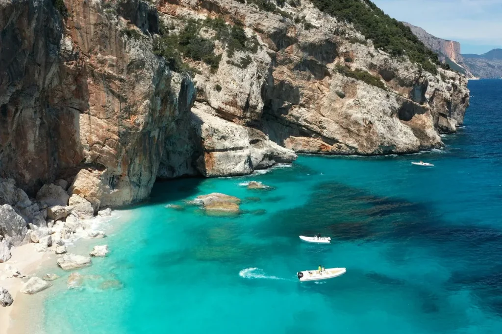 Solo travel sardinia: turquoise water and a white beach in a hidden cove on Sardinia's Gulf of Orosei, ringed by tall white limestone cliffs, with a few small boats anchored in the shallows.