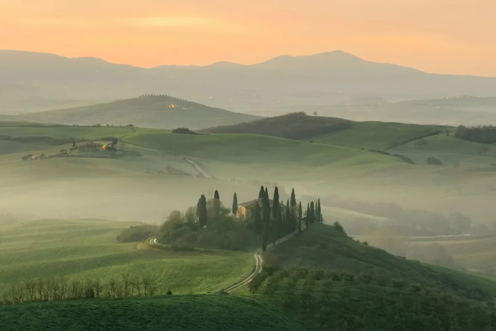 A stone farmhouse ringed by cypress trees on a misty green hill in the Val d'Orcia at dawn, one of the best day trips from Florence for women over 50