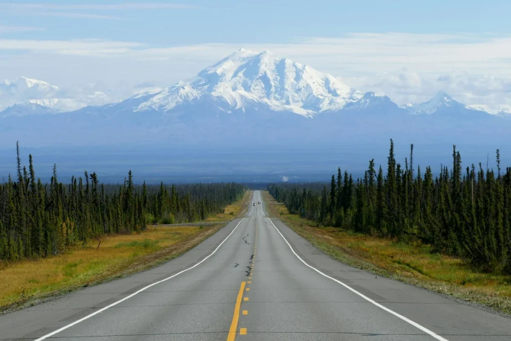 Solo travel Alaska: an open highway leading toward a snow-capped mountain through spruce forest under a blue sky, a guide for women over 50.
